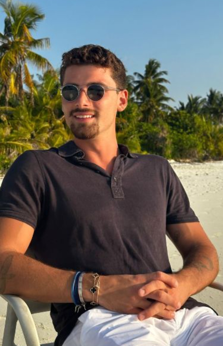 Iman Gadzhi smiling on a tropical beach, wearing sunglasses and a navy polo shirt.